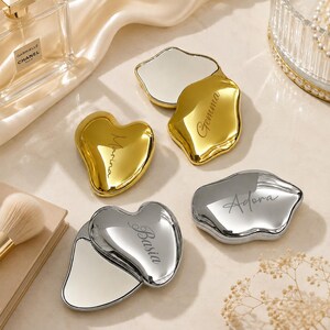 May include: Four compact mirrors in heart and irregular cloud shapes with metallic gold and silver finishes. Each mirror features a custom engraved name: Marina, Gemma, Adora, and Basia. The mirrors are shown in both closed and open positions.