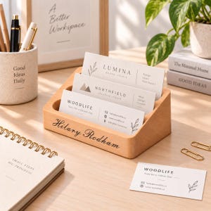 Puede incluir: Un tarjetero de madera de color claro con tres ranuras escalonadas, personalizado con el nombre Hilary Rodham en letra cursiva negra. El soporte contiene tres tarjetas de visita blancas con texto negro e ilustraciones botánicas lineales.