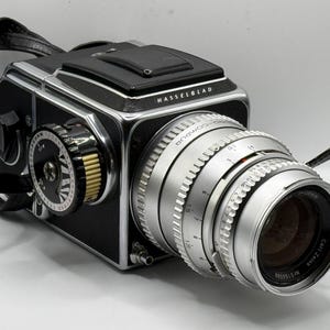 May include: A black and silver Hasselblad medium format camera with a Carl Zeiss lens. The camera features a silver Compur shutter lens barrel with visible aperture and focus markings, a black leatherette body, and a black camera strap.
