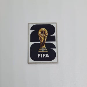 May include: A rectangular metal pin featuring a black and white design with the large black numbers 26. Centered over the numbers is a gold-colored trophy graphic. The word FIFA is printed in white capital letters at the bottom of the pin.