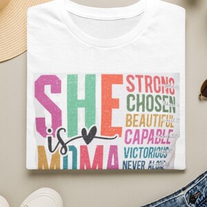 May include: A white t-shirt featuring a colorful graphic print with the text "SHE is MOMA" in large block letters, accompanied by a list of words including "STRONG," "CHOSEN," "BEAUTIFUL," "CAPABLE," "VICTORIOUS," and "NEVER ALONE" in various colors.