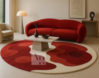 May include: A red, cream, and burgundy area rug with an abstract floral design. The rug is circular and features a layered pattern of varying shades of red and cream. The rug is placed in a living room setting with a red sofa and a light-colored coffee table.