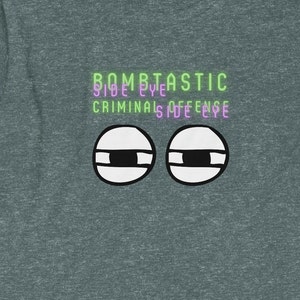 May include: A dark heather grey t-shirt with the text "BOMBTASTIC SIDE EYE CRIMINAL SIDE EYE" in green and purple. The shirt features a cartoon illustration of two eyes with black pupils and white sclera.