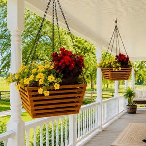 May include: Two square wooden hanging planters with horizontal slats suspended by black metal chains. Each planter is filled with vibrant yellow flowers and deep red blossoms, creating a colorful display on a white porch with railings.