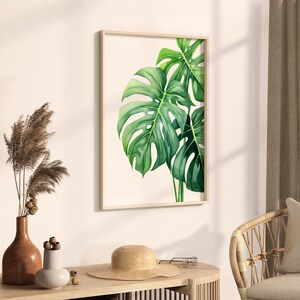 May include: A framed watercolour painting of green monstera leaves on a white background. The painting is hanging on a white wall in a room with a wicker chair and a vase of dried flowers.