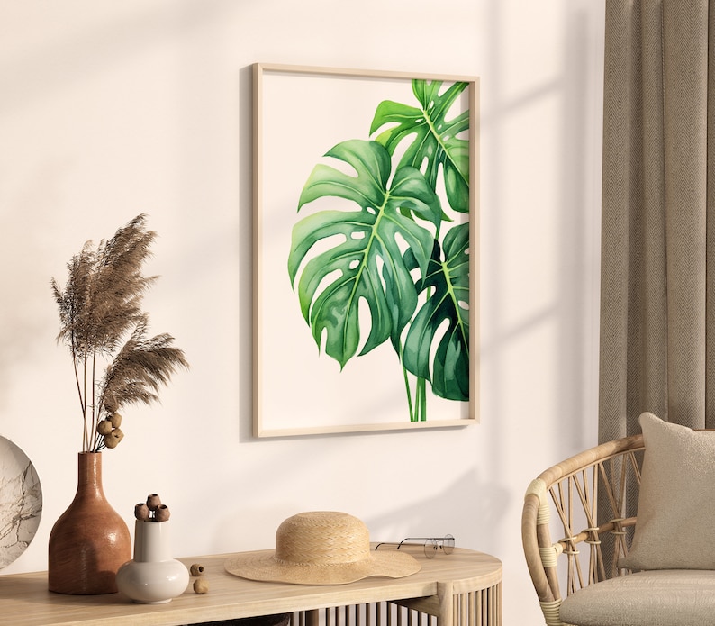 May include: A framed watercolour painting of green monstera leaves on a white background. The painting is hanging on a white wall in a room with a wicker chair and a vase of dried flowers.