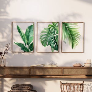 May include: Three framed botanical prints featuring different types of tropical leaves. The prints are in a modern style with a white background and green leaves. The prints are hung on a white wall above a wooden console table.