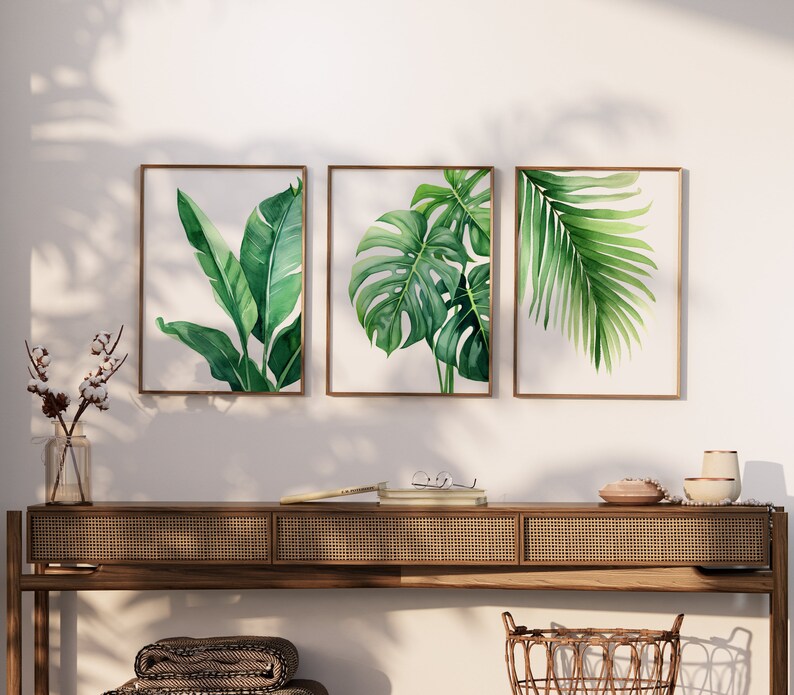 May include: Three framed botanical prints featuring different types of tropical leaves. The prints are in a modern style with a white background and green leaves. The prints are hung on a white wall above a wooden console table.