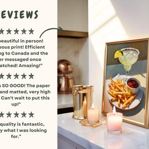 May include: A framed print of a painting depicting a margarita cocktail with a lime wedge, french fries, and a small dish of ketchup. The print is displayed on a white countertop with two candles and a metal ice bucket.