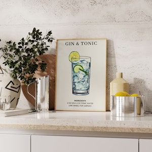 May include: A framed print with a white background and the text "Gin & Tonic" in black. The print features a hand-drawn illustration of a glass of gin and tonic with a lime wedge and ice cubes. The text "Ingredients" is below the illustration, followed by a list of ingredients: "2 oz Gin + 4 oz Tonic Water, Lime Wheel for Garnish."