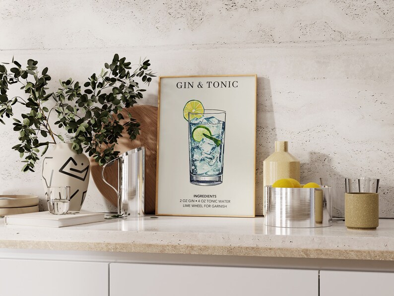 May include: A framed print with a white background and the text "Gin & Tonic" in black. The print features a hand-drawn illustration of a glass of gin and tonic with a lime wedge and ice cubes. The text "Ingredients" is below the illustration, followed by a list of ingredients: "2 oz Gin + 4 oz Tonic Water, Lime Wheel for Garnish."