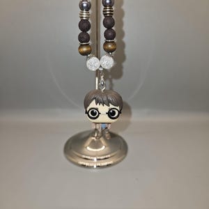 May include: A necklace featuring a Harry Potter Funko Pop charm. The necklace has a beaded design with various colors and textures, including dark gray, brown, and clear beads. The charm is a miniature figure of Harry Potter wearing glasses.