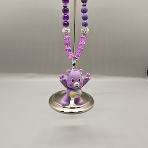 May include: A purple necklace featuring a Care Bear pendant with a rainbow belly. The necklace has a beaded design with purple and clear beads. The pendant is a light purple color and is standing on a silver stand.