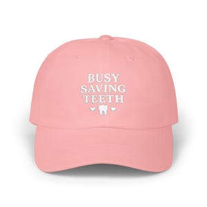 May include: A light pink baseball cap with the embroidered text "BUSY SAVING TEETH" in white. The design includes a tooth and two hearts. The cap is a classic six-panel design with a curved brim.