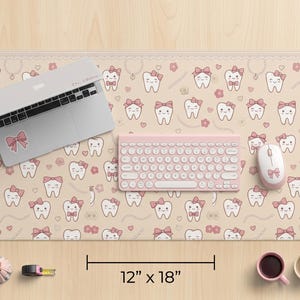 Peut inclure: Tapis de bureau beige avec un motif répété de dents de dessin animé avec des nœuds roses et des fleurs. Un ordinateur portable, un clavier et une souris roses sont posés dessus. Le tapis mesure 30 cm x 46 cm.