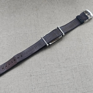 May include: A dark brown leather watch strap with a silver-colored buckle and keepers. The strap has several holes for adjustment and is designed to fit a watch. The strap is approximately 9 inches long.