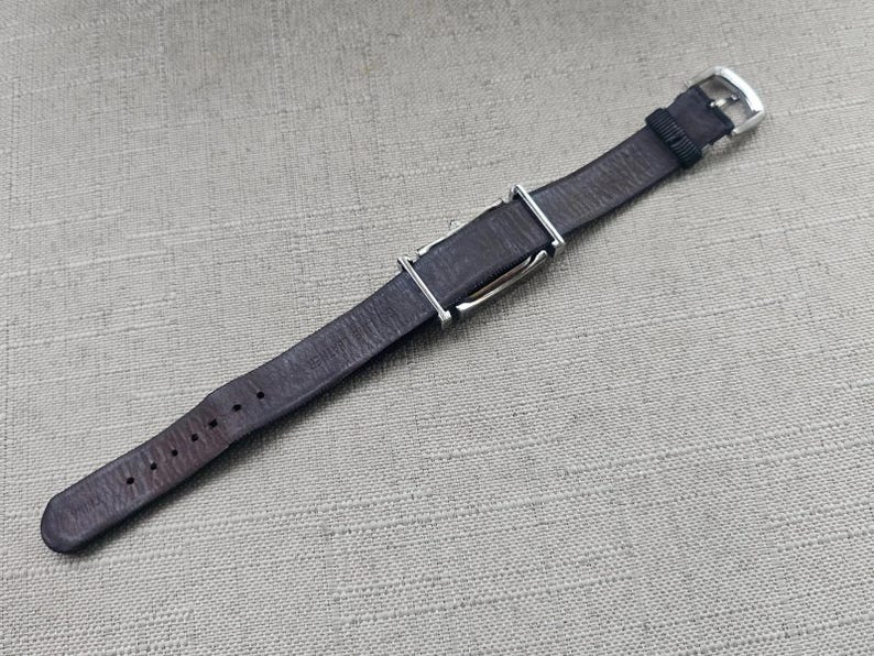 May include: A dark brown leather watch strap with a silver-colored buckle and keepers. The strap has several holes for adjustment and is designed to fit a watch. The strap is approximately 9 inches long.