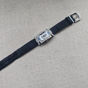 May include: A rectangular wristwatch with a black fabric band and a silver buckle. The watch face is rectangular with a silver frame and small blue accents. The watch is adorned with small, clear gemstones.