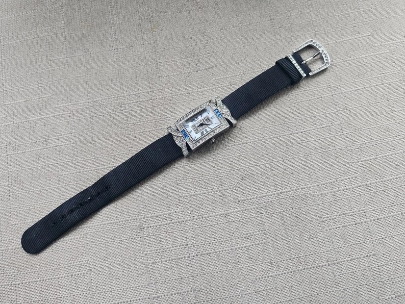 May include: A rectangular wristwatch with a black fabric band and a silver buckle. The watch face is rectangular with a silver frame and small blue accents. The watch is adorned with small, clear gemstones.