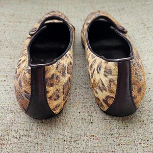 May include: A pair of flat shoes with a leopard print pattern. The shoes have a brown and tan colour scheme with a dark brown trim and a strap across the top. The soles are black.