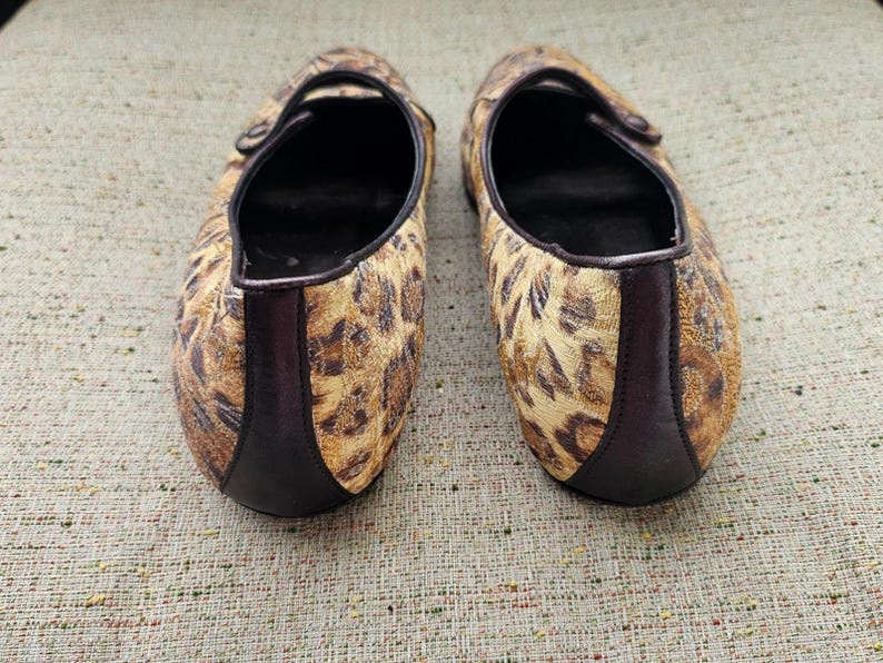 May include: A pair of flat shoes with a leopard print pattern. The shoes have a brown and tan colour scheme with a dark brown trim and a strap across the top. The soles are black.