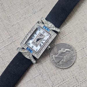 May include: A rectangular wristwatch with a white face, Roman numerals, and a black band. The watch is adorned with small diamonds and blue accents. A quarter coin is next to the watch.