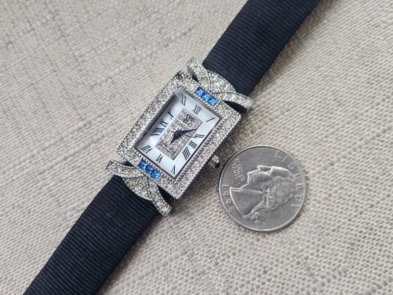 May include: A rectangular wristwatch with a white face, Roman numerals, and a black band. The watch is adorned with small diamonds and blue accents. A quarter coin is next to the watch.