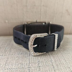 May include: Close-up of a wristwatch with a navy blue ribbed band and a silver buckle. The buckle is adorned with small, clear crystals. The watch face is visible in the background.