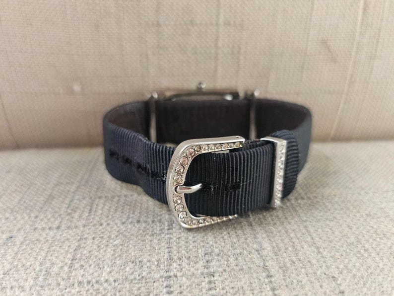 May include: Close-up of a wristwatch with a navy blue ribbed band and a silver buckle. The buckle is adorned with small, clear crystals. The watch face is visible in the background.