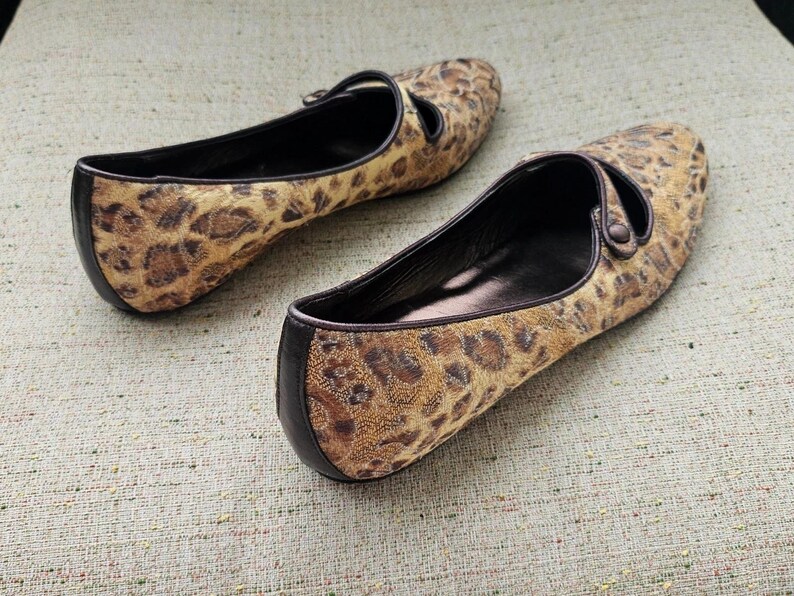 May include: A pair of leopard print ballet flats. The shoes feature a brown and tan spotted pattern with black trim and a strap. The shoes are made of a soft, textured material.