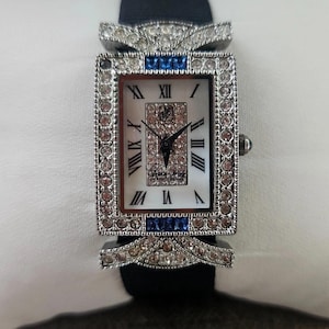 May include: A rectangular wristwatch with a silver-colored bezel encrusted with clear and pink crystals. The watch face is white with black Roman numerals and black hands. Blue crystals accent the top and bottom of the bezel. The watch has a black band.