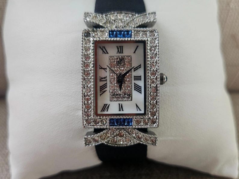 May include: A rectangular wristwatch with a silver-colored bezel encrusted with clear and pink crystals. The watch face is white with black Roman numerals and black hands. Blue crystals accent the top and bottom of the bezel. The watch has a black band.