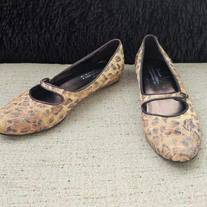 May include: A pair of women's flats with a brown and tan animal print pattern. The shoes have a Mary Jane strap and a dark brown trim. The flats are displayed on a light beige textured surface.