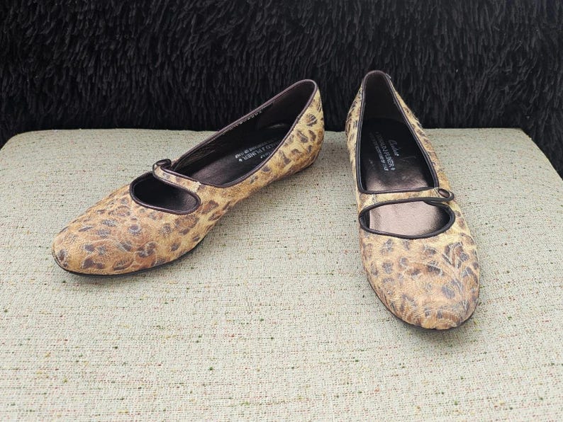 May include: A pair of women's flats with a brown and tan animal print pattern. The shoes have a Mary Jane strap and a dark brown trim. The flats are displayed on a light beige textured surface.