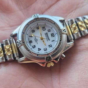 Puede incluir: Un reloj de pulsera Sharp Opti-Glo plateado y dorado con esfera blanca. El reloj tiene una esfera redonda con números negros y agujas doradas. La correa es una combinación de eslabones metálicos plateados y dorados.