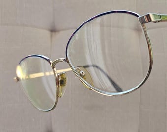 Peut inclure: Une paire de lunettes avec une monture en métal doré et des verres transparents. Les verres sont de forme arrondie. La monture a un design fin et délicat. Les lunettes sont posées sur un fond neutre.