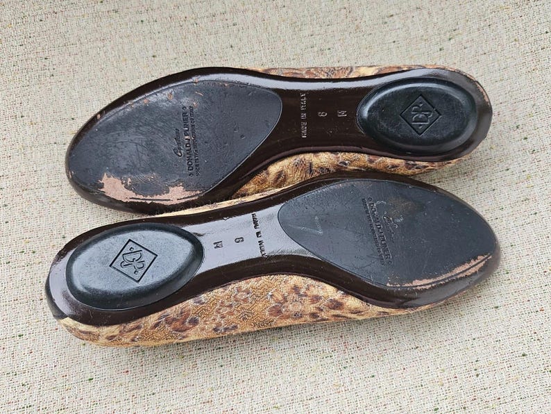 May include: A pair of women's flats with a leopard print design. The soles are dark brown with a black heel pad and the brand name "Donald J Pliner" is visible. The shoes are size 9 M.