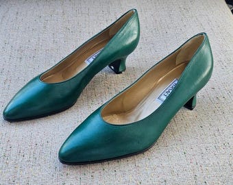 May include: A pair of emerald green leather pumps with pointed toes and small heels. The shoes have a classic design and a label inside. The pumps are displayed on a neutral-toned surface.