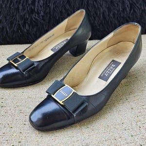 Può includere: Un paio di décolleté in pelle nera con punta quadrata e tacco piccolo. Ogni scarpa presenta un fiocco nero con fibbia color oro. Il marchio "BALLY" è visibile sulla soletta.