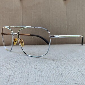 May include: Silver-toned metal eyeglasses featuring a double bridge design with a textured top bar. The frames include clear lenses, gold-toned adjustable nose pads, and thin temples with dark-colored tips, showcasing a classic aviator-inspired shape.