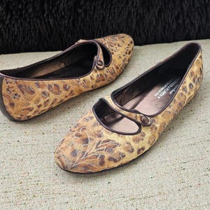 May include: A pair of women's flats featuring a tan and brown leopard print design. The shoes have a rounded toe and a strap. The soles and trim are dark brown. The shoes are made of a patterned fabric.