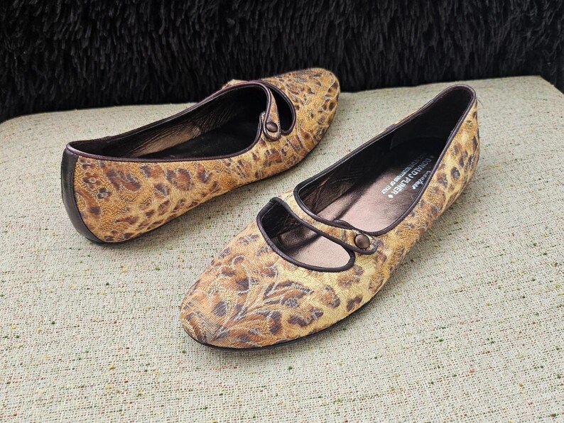 May include: A pair of women's flats featuring a tan and brown leopard print design. The shoes have a rounded toe and a strap. The soles and trim are dark brown. The shoes are made of a patterned fabric.