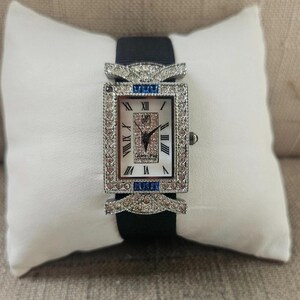 May include: A rectangular wristwatch with a white face and Roman numeral hour markers. The watch features a silver-colored bezel and is adorned with small, sparkling stones and blue accents. It has a black band and is displayed on a white pillow.