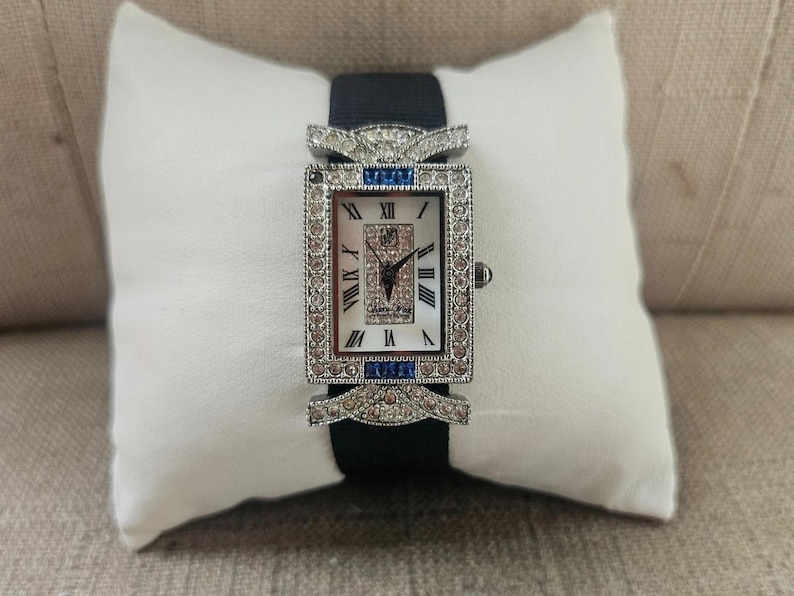 May include: A rectangular wristwatch with a white face and Roman numeral hour markers. The watch features a silver-colored bezel and is adorned with small, sparkling stones and blue accents. It has a black band and is displayed on a white pillow.