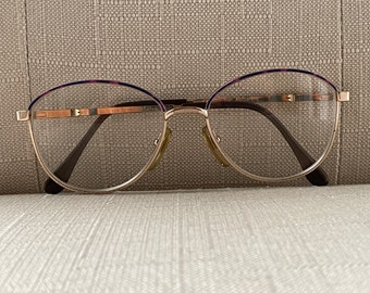 May include: Gold-rimmed glasses with a purple and brown tortoiseshell frame. The glasses have a clear lens and the text "135 02 51-18 140" is printed on the right arm.