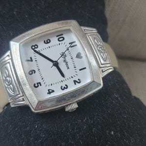 Könnte beinhalten: Eine silberfarbene Brighton-Uhr mit rechteckigem Zifferblatt und weißem Zifferblatt. Die Uhr hat schwarze Ziffern, Zeiger und das Brighton-Logo. Sie hat ein helles Lederarmband und dekorative Akzente an den Seiten.