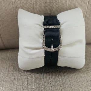 May include: A black watch band with a silver buckle, adorned with small, sparkling embellishments, is displayed on a white, rectangular pillow. The pillow is resting on a textured, light brown surface.