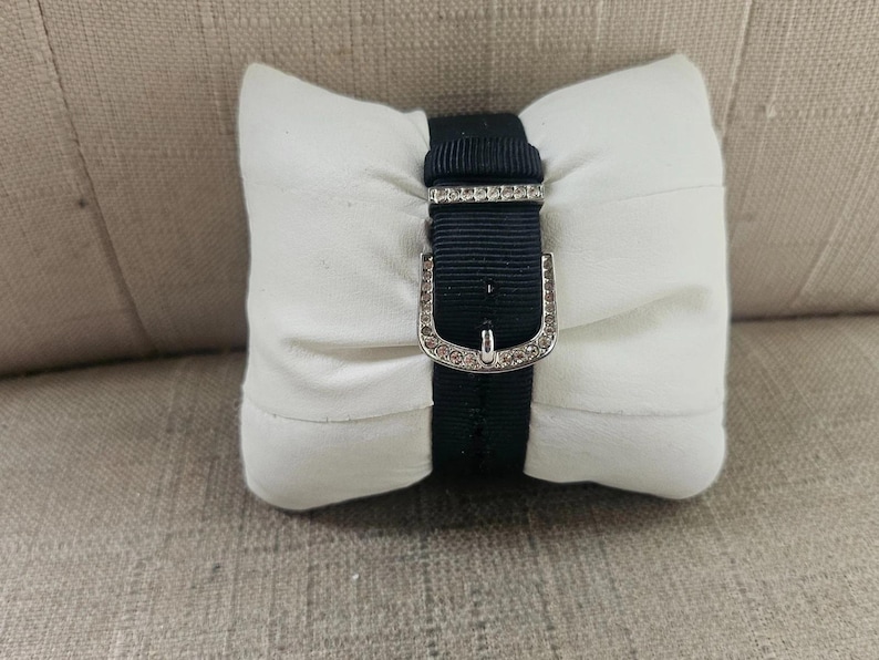 May include: A black watch band with a silver buckle, adorned with small, sparkling embellishments, is displayed on a white, rectangular pillow. The pillow is resting on a textured, light brown surface.