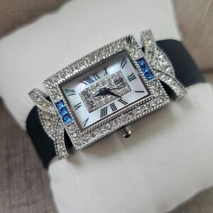 May include: A rectangular wristwatch with a silver-colored bezel encrusted with clear crystals. The watch face has Roman numerals and a mother-of-pearl background. Blue crystals accent the sides, and a black band completes the design.