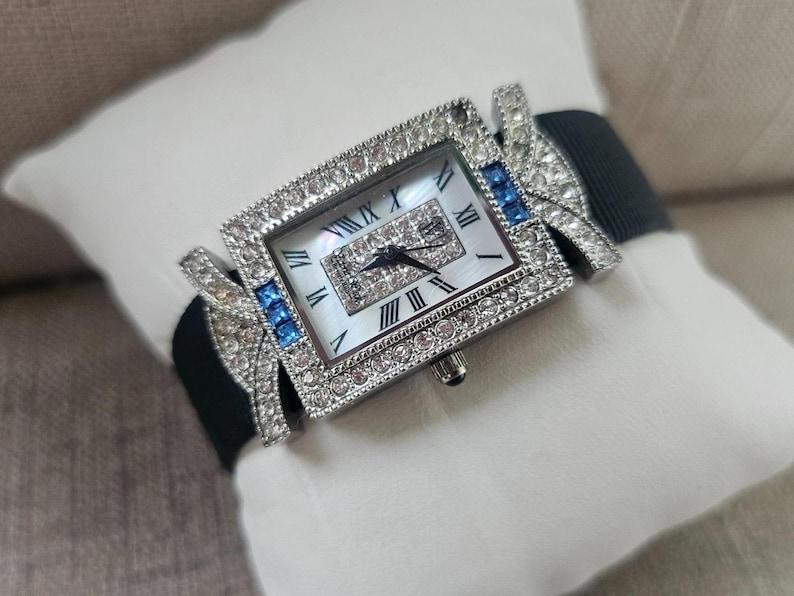 May include: A rectangular wristwatch with a silver-colored bezel encrusted with clear crystals. The watch face has Roman numerals and a mother-of-pearl background. Blue crystals accent the sides, and a black band completes the design.